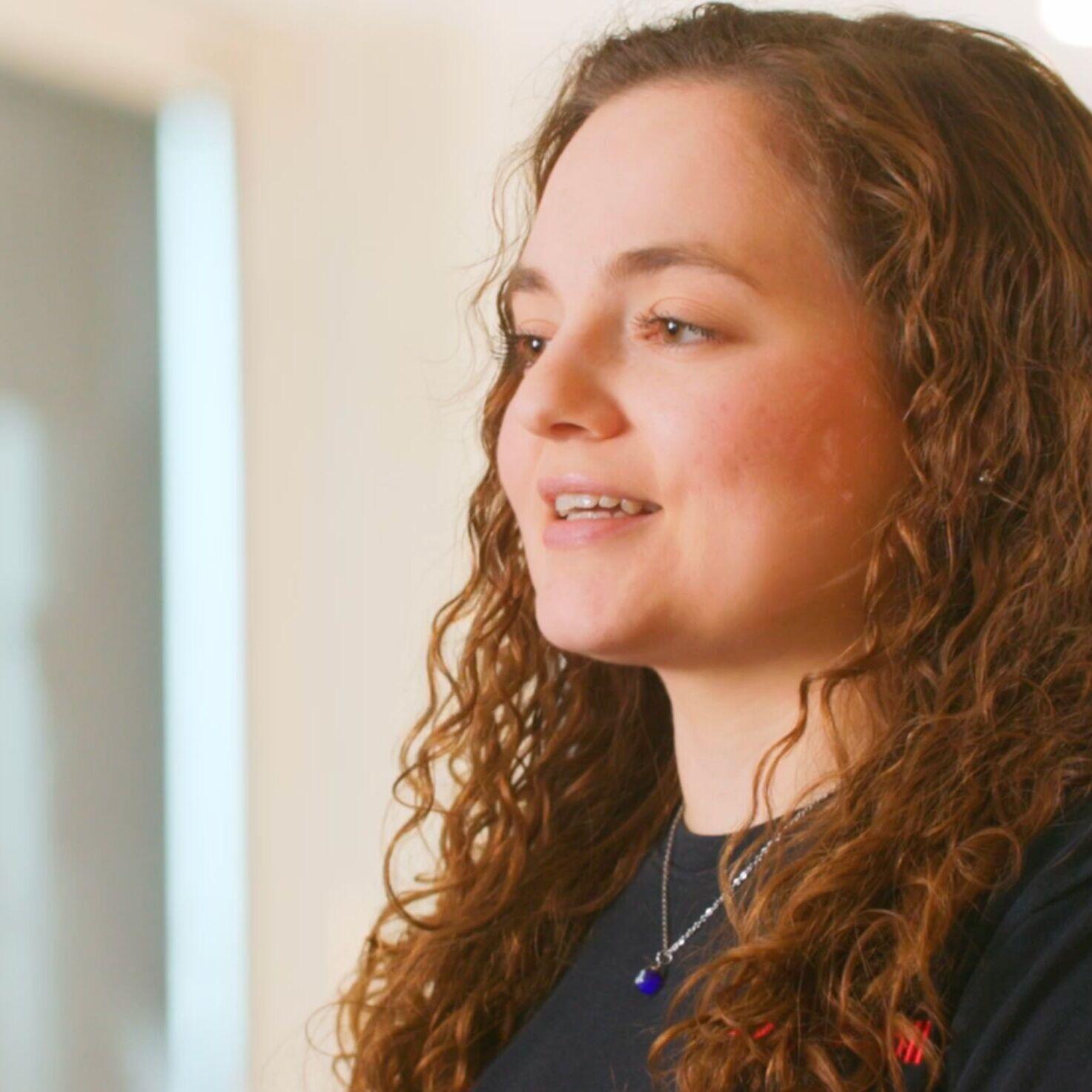 A person with long, curly brown hair standing indoors.