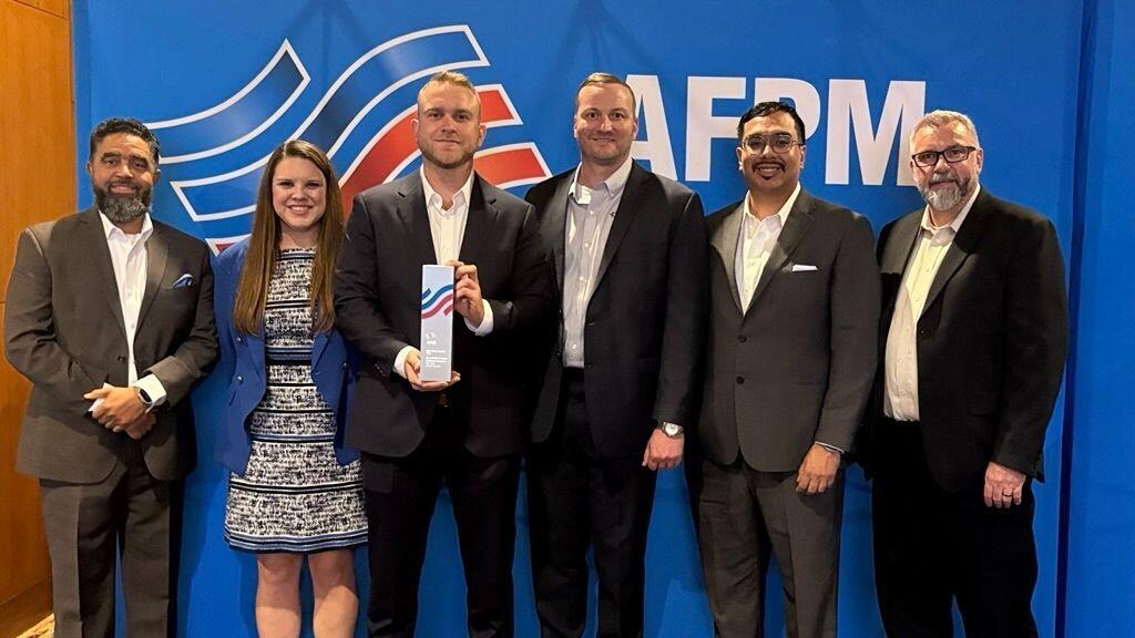 A group of six people in business attire pose for a photo in front of a blue backdrop with the AFPM logo. The person in the center holds a trophy with a wavy red-and-blue design.