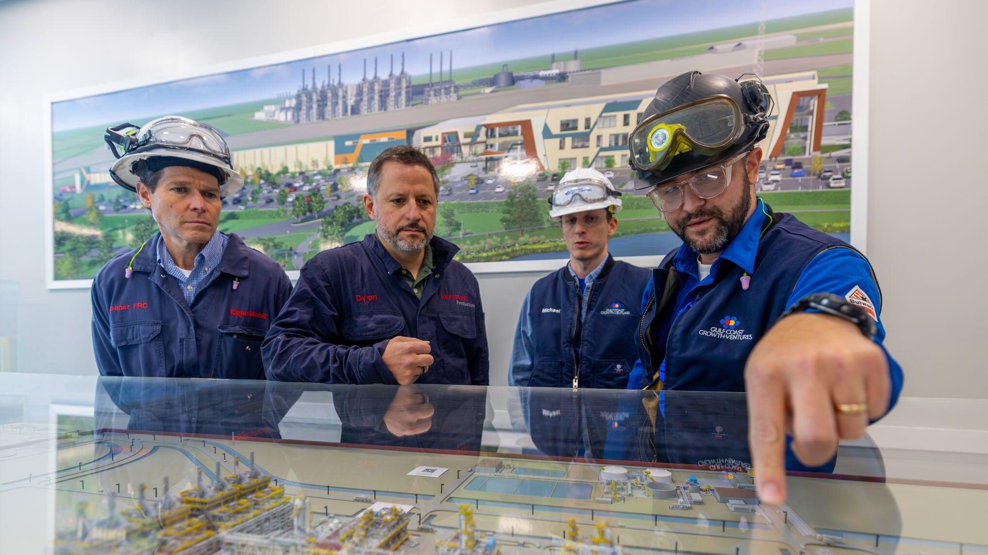 Four workers wearing protective gear stand around a glass-covered scale model of an industrial plant.