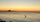 A serene sunset scene over a calm bay shows the sky fading from orange near the horizon to pale blue above. In the foreground, dark rocks line the shore while gentle ripples move across the water. A lone wading bird, likely a heron or egret, stands in the shallow water, its reflection visible. Far in the distance, a pier and a distinctive structure are silhouetted against the glowing horizon.