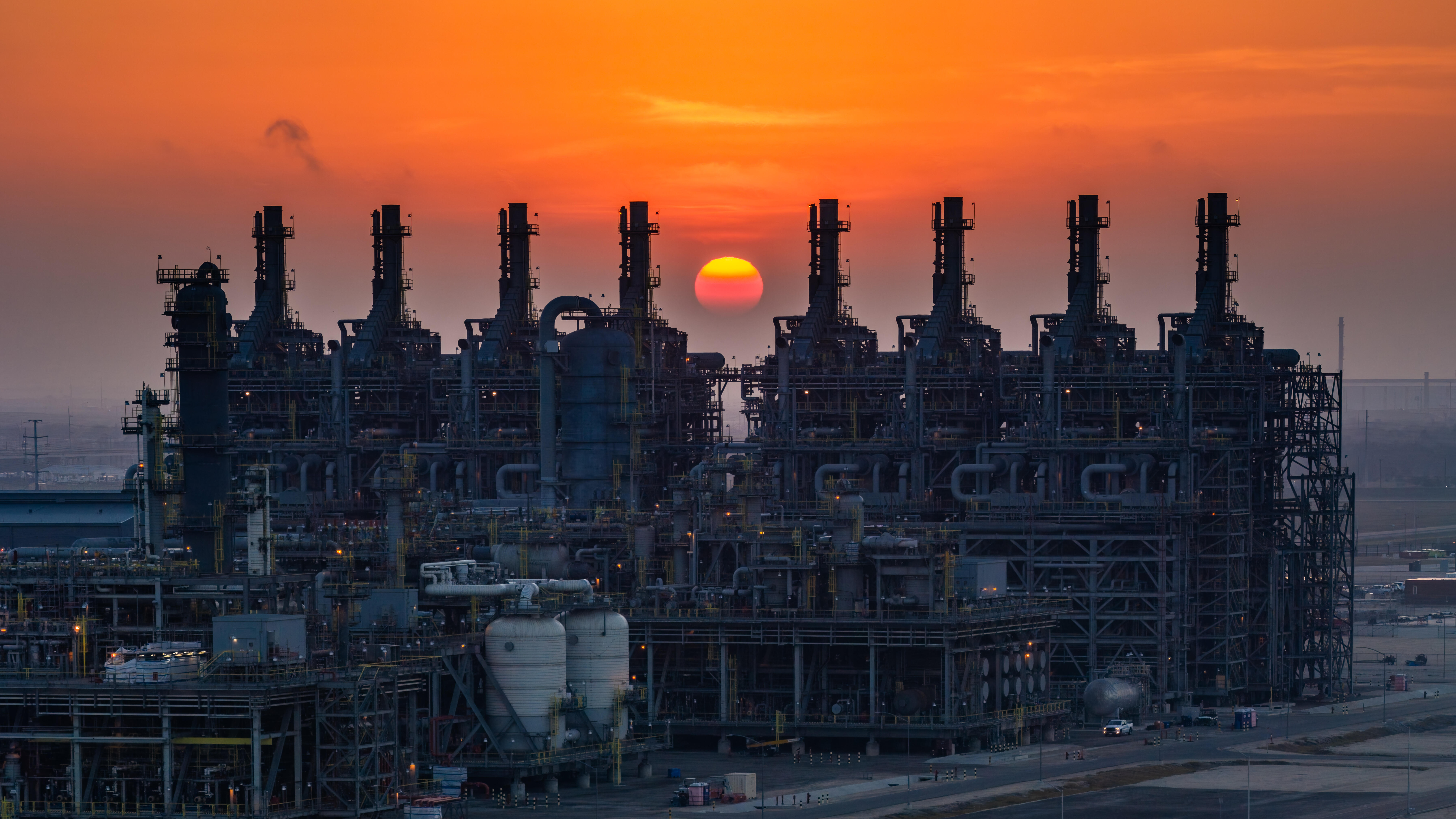 An expansive industrial refinery complex is silhouetted against a vivid orange sunset.