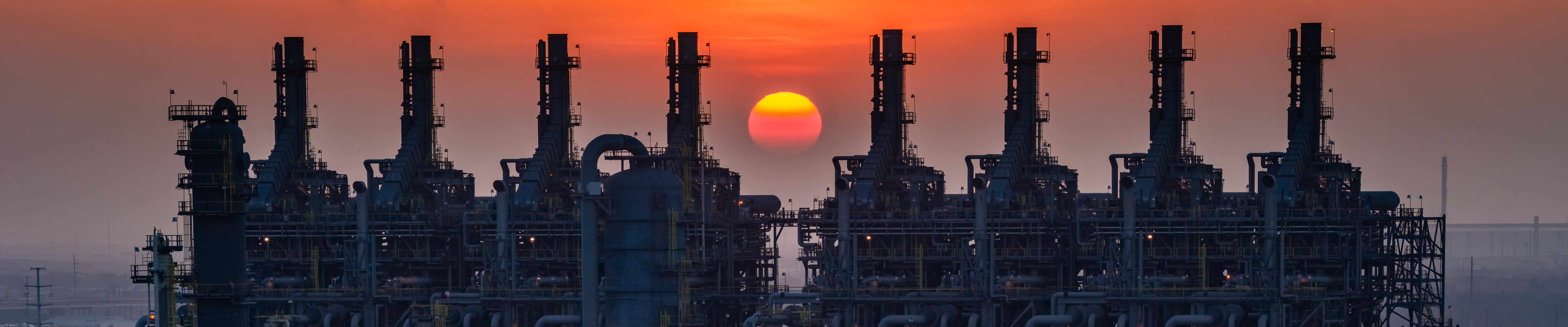 An expansive industrial refinery complex is silhouetted against a vivid orange sunset.