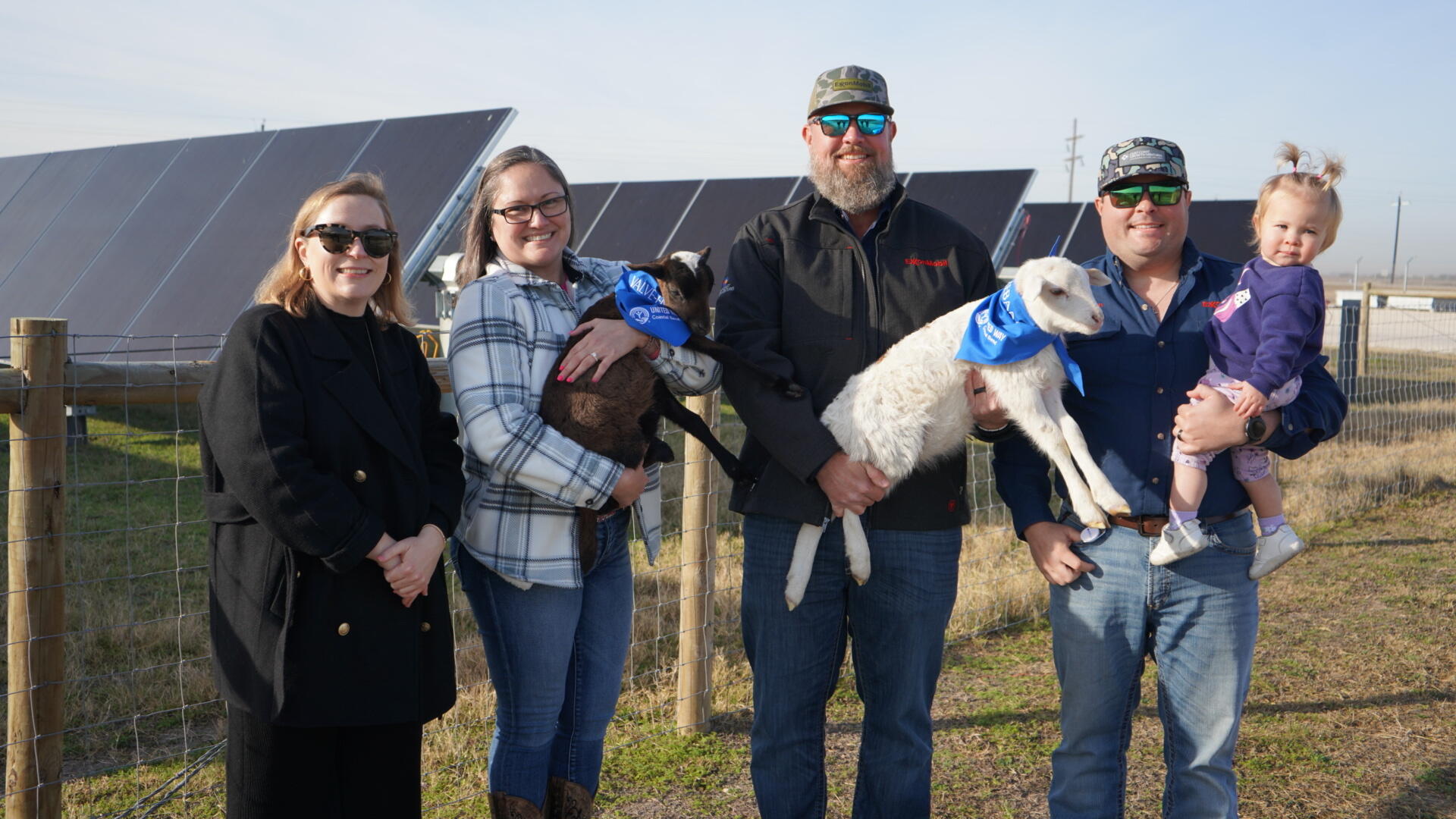 Five adults and a toddler stand together outdoors in front of rows of solar panels and a wire fence. Two of the adults are holding small goats wearing blue bandanas, and the man on the right is holding the child.