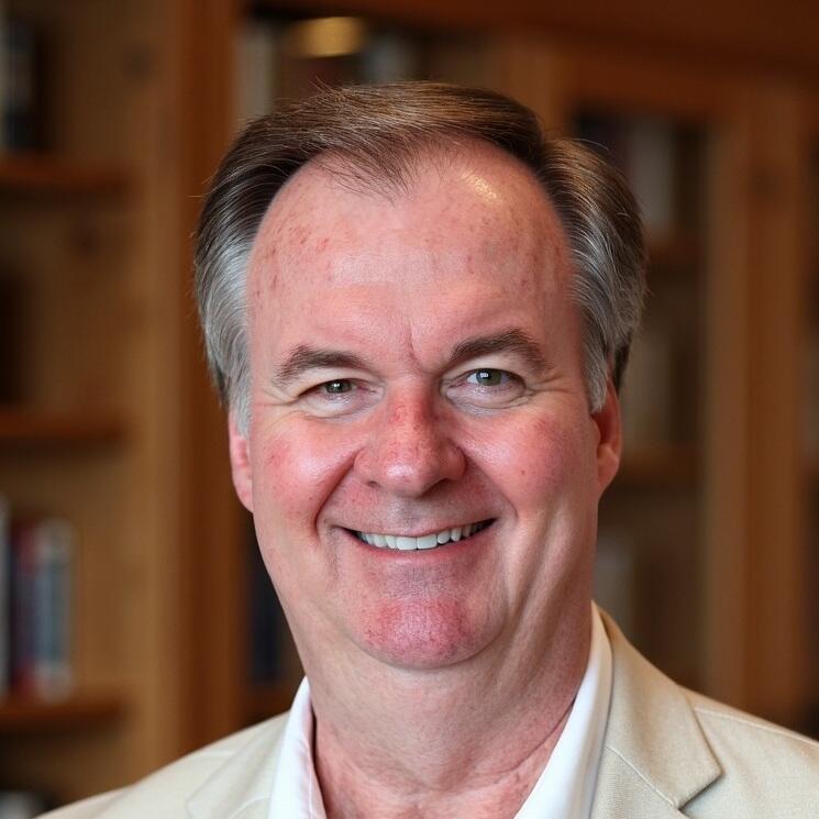 A man is smiling in a warmly lit indoor setting, possibly an office or library. He has short, graying hair and is wearing a light beige blazer over a white dress shirt.