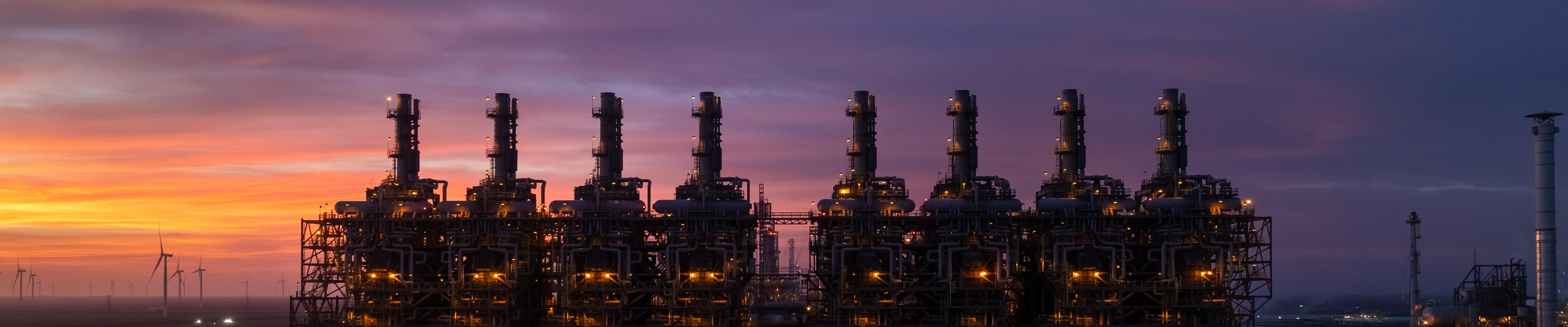 A massive industrial complex is lit up at dusk, with rows of tall stacks and intricate piping glowing amber against a purple and blue sky.