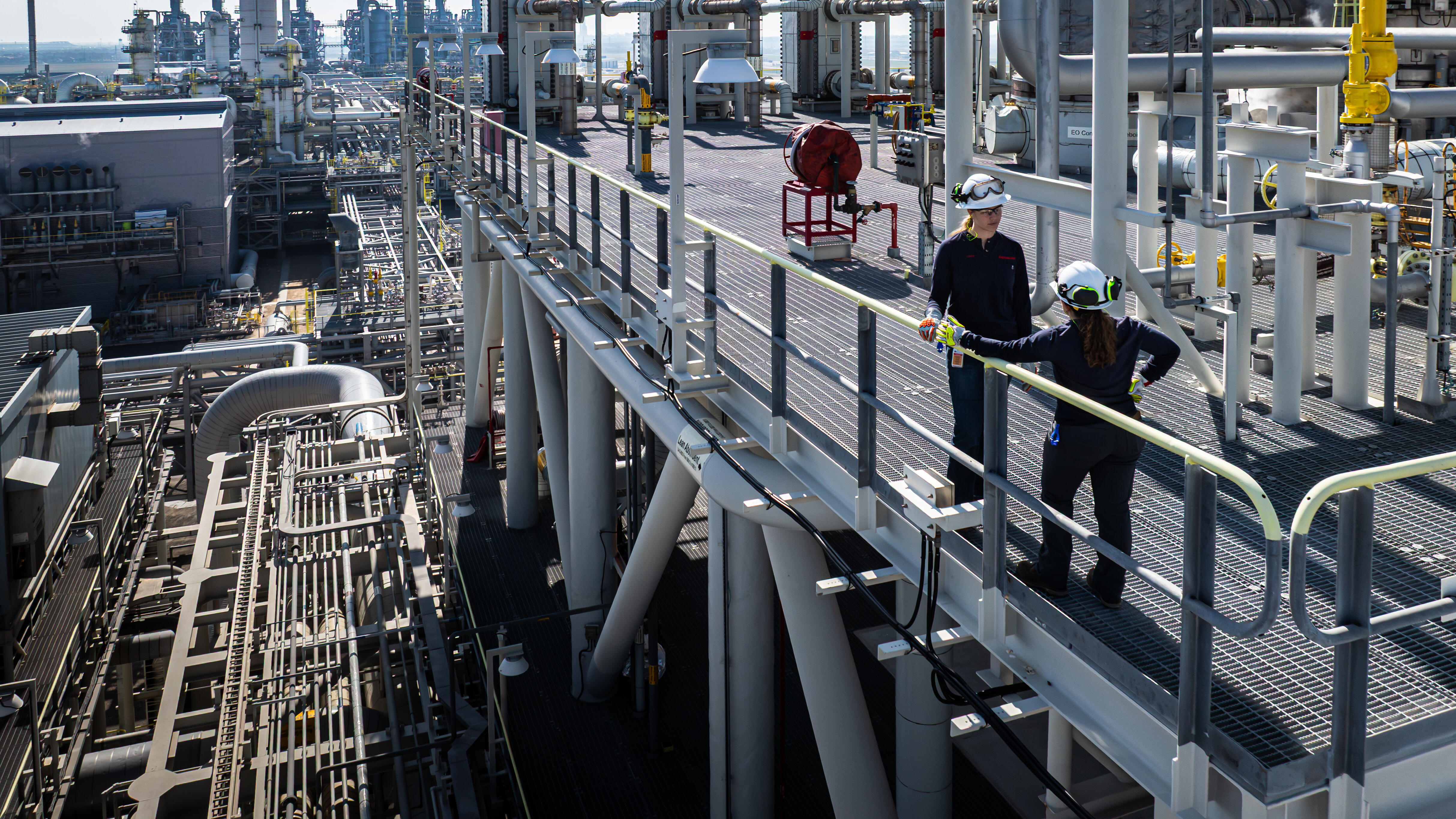 An expansive industrial plant with numerous pipes, towers, and metal structures stretching into the distance. Two workers in safety gear—helmets, goggles, gloves, and radios—stand on an elevated grated platform, talking near a handrail.