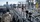 An expansive industrial plant with numerous pipes, towers, and metal structures stretching into the distance. Two workers in safety gear—helmets, goggles, gloves, and radios—stand on an elevated grated platform, talking near a handrail.