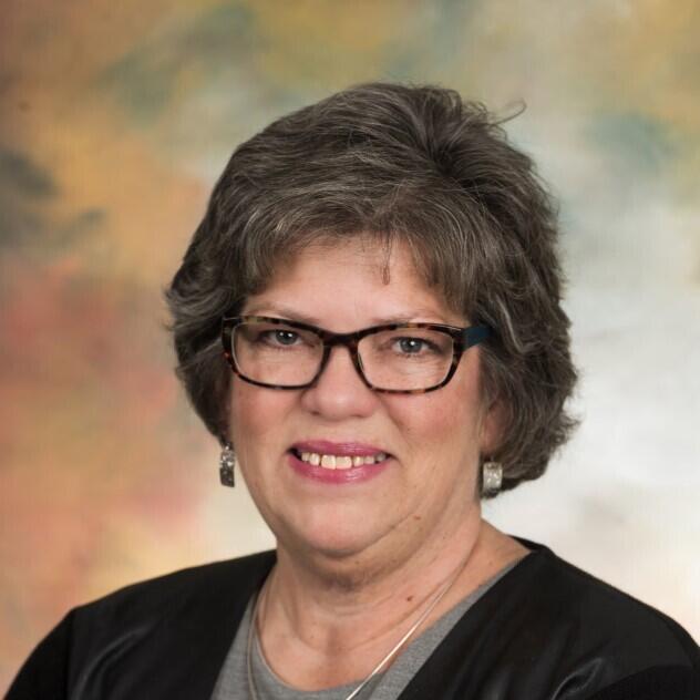 A professional portrait of woman with short, wavy gray hair and glasses.