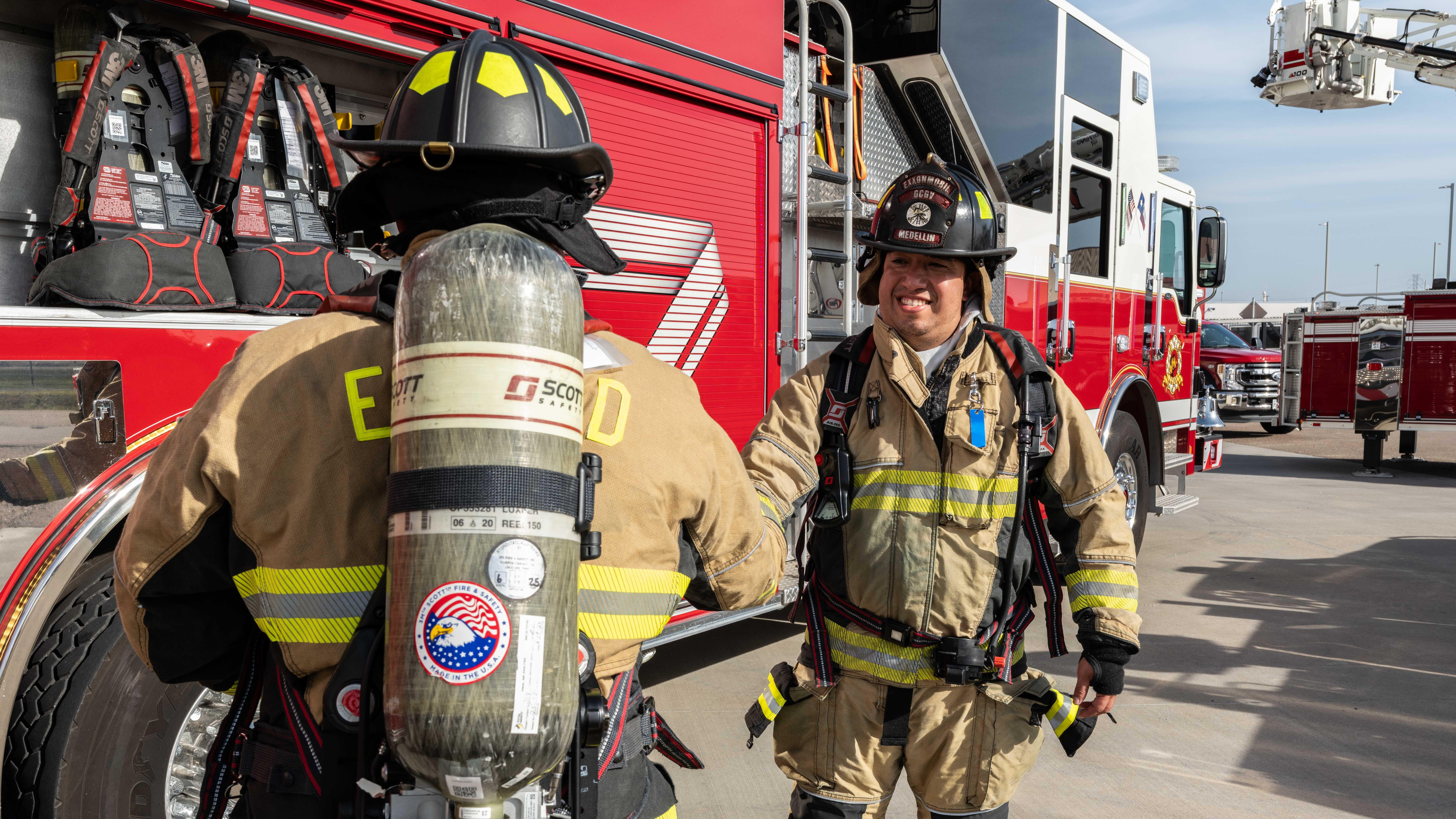 Two firefighters in full turnout gear stand beside a bright red fire engine labeled “ENGINE 12.”