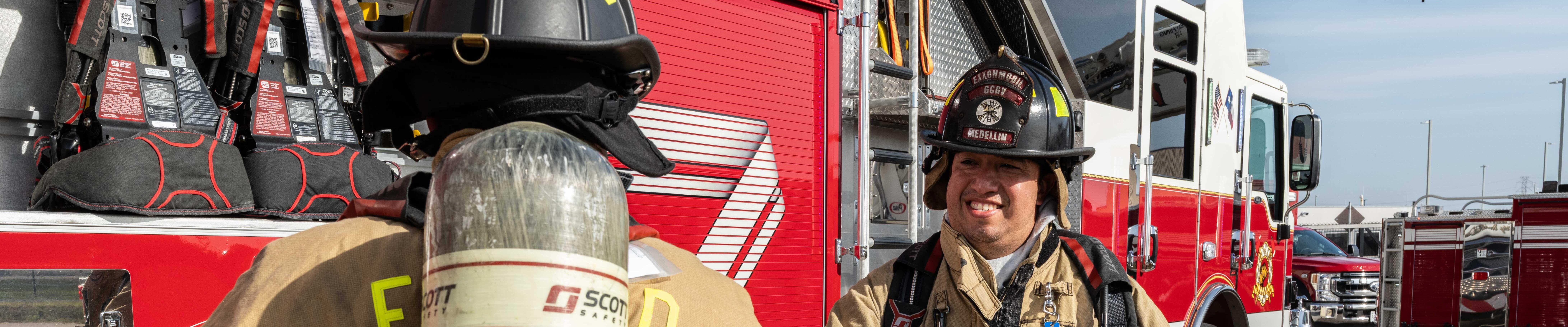 Two firefighters in full turnout gear stand beside a bright red fire engine labeled “ENGINE 12.”
