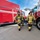 Four firefighters in full turnout gear are walking between two red fire trucks.