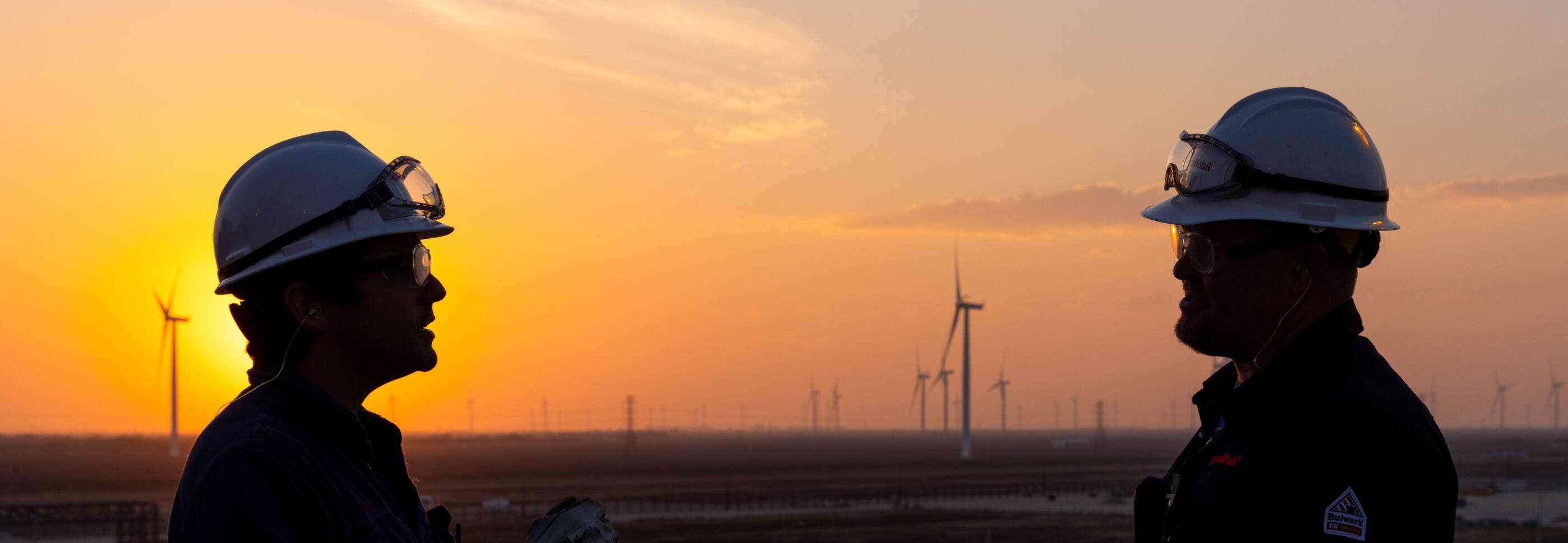 Two employees wearing hard hats and safety goggles with sunset in the background.